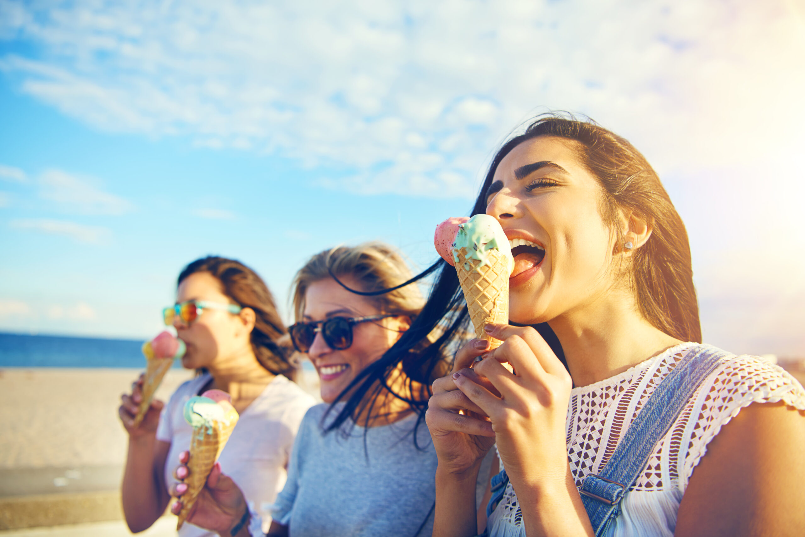 Three young woman eating ice cream cones at the seaside as they stroll along a waterfront promenade on a hot summer day during their vacation
